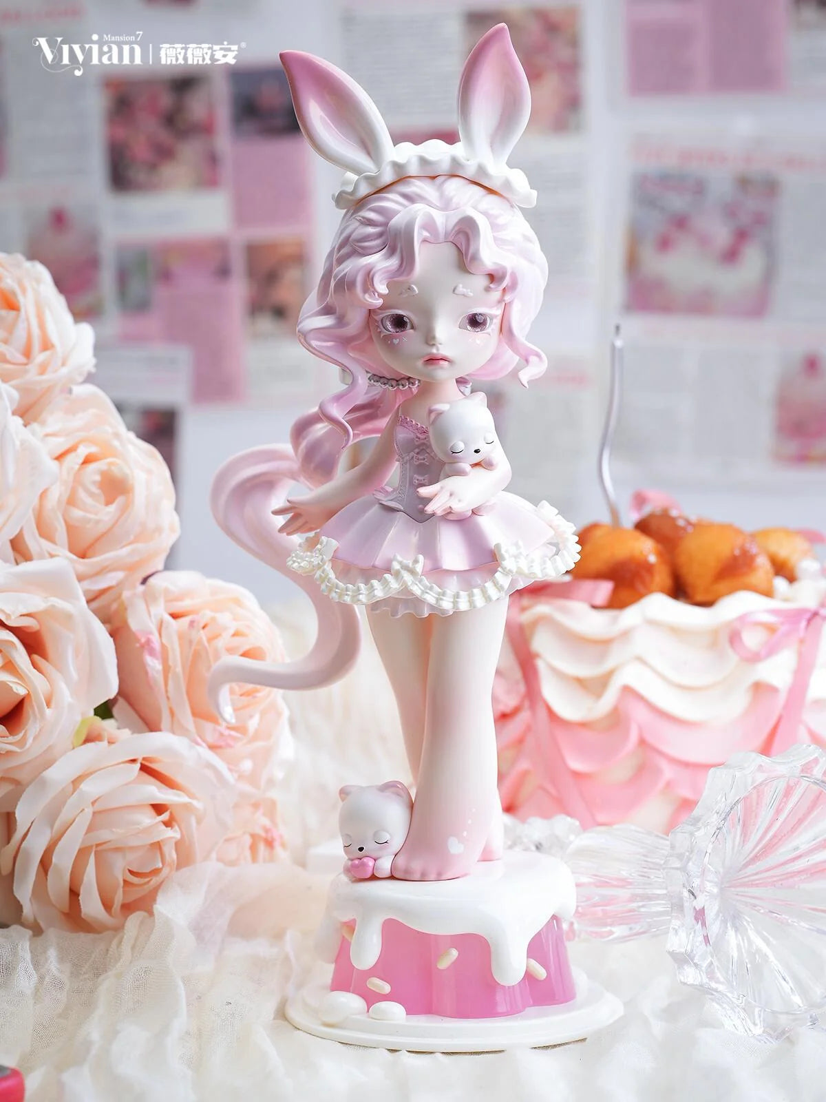 A pink bunny-themed figurine of a girl with long pink hair, bunny ears, and a tutu holding a small white bunny, displayed on a decorated cake base with roses and a pink dessert in the background.